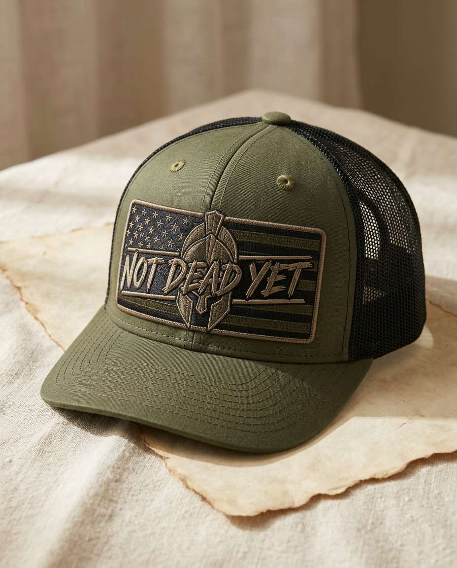Olive green trucker hat with black mesh back and an embroidered Not Dead Yet spartan helmet flag patch